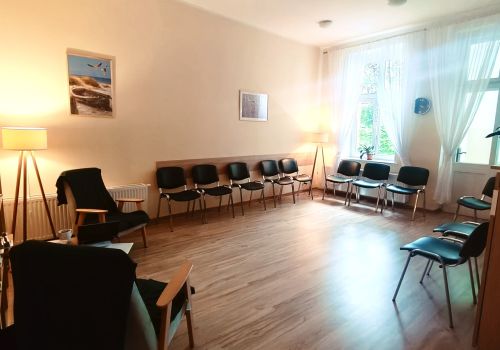 Kolejne zdjęcie pomieszczenia terapii grupowej, wykonane pod innym kątem - na zdjęciu widać ustawione w kwadracie krzesła, lampę stojącą, rzucającą nastrojowe światło. Na jednej ścianie wiszą oprawione zdjęcia sopockie, przestrzeń drugiej w większości wypełniają okna.