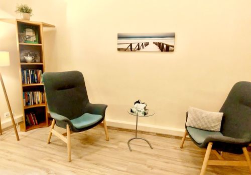 Zdjęcie pomieszczenia terapii indywidualnej. Na zdjęciu widać dwa wygodne fotele, regał z książkami i niewielki stolik, na którym umieszczono pudełko z chusteczkami. Na ścianie wisi obraz prezentujący sopockie molo.