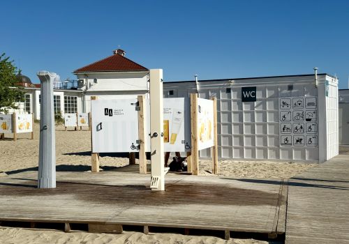 Zdjęcie urządzonego odcinka plaży dostosowanego do potrzeb osób ze specjalnymi potrzebami. Na zdjęciu widać ułożony na piasku podest doprowadzający do przebieralni, prysznica i toalet.